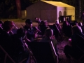 Noc, Ludzie siedzący na leżakach na terenie festiwalu, oświetleni fioletowym światłem telebimu. W tle biały namiot.